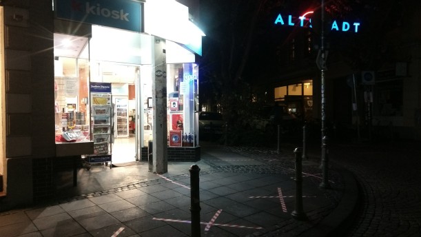 Nachtansicht eines erleuchteten Kiosk an einer menschenleeren Straßenecke, im Hintergrund leuchtet ein Schriftzug "Altstadt".