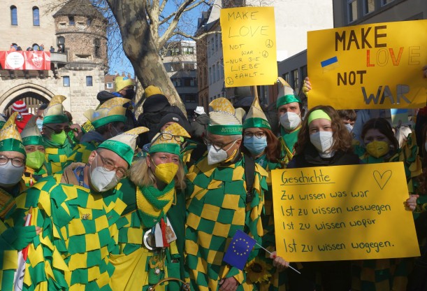 Eine Menschengruppe in grün-gelben Lappenclownkostümen, einige halten Plakate mit Friedensbotschaften