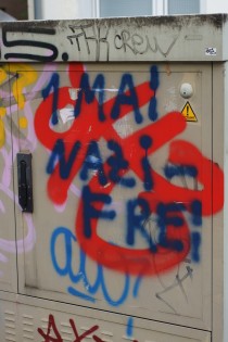 Trafokasten mit mehrfarbigen Graffiti. Über roten Ornamenten ist mit blauer Schrift der Slogan „1.Mail Nazi-Frei“ gesprayt.