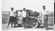 Ein Mann und zwei Frauen stehen um ein kleines Auto, auf das ein Koffer geschnallt ist. Die Frauen tragen knielange Röcke und weiße, ärmellose Blusen, der Mann Hose und langärmeliges Hemd.