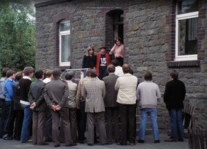 Eine Gruppe Männer vor einer Haustür an der drei junge Frauen stehen.
