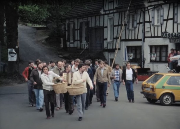 Junge Männer gehen durch einen Dorfstraße. Manche von ihnen tragen Körbe.
