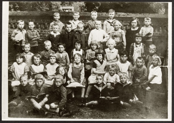 Eine Schulklasse posiert für das Foto mit ihrer Lehrerin auf dem Schulhof. Manche Kinder haben Bücher im Schoß liegen.