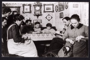 Eine Gruppe von Menschen sitzt in einer Wohnstube um einen Tisch herum. Die Frau rechts im Bild sitzt am Spinnrad und ist, ebenso wie die Frau ganz links im Bild, mit Handarbeit beschäftigt, neben ihr ein Mann der in einem Buch liest und Pfeife raucht, am Tisch sitzen Kinder und Erwachsene vor einem Spielbrett. In der Tür zur Stube stehen drei weitere Menschen, die das Spiel beobachten.