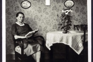 Eine Frau sitzt auf einem Sessel an einem Tisch und liest ein Buch. Sie trägt ein gepunktetes Sommerkleid mit kurzen Puffärmeln und weißer Ausschnitteinfassung. Auf dem runden Tisch neben ihr steht eine Topfpflanze, an der mit einer geblümten Tapete tapezierten Wand hängen kleine Bilder.