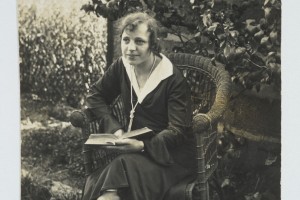 Porträt einer Frau, sitzend auf einem Korbstuhl mit geöffnetem Buch im Schoß.