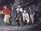 Gruppenbild von Bergarbeitern in einem Stollen. Die Arbeiter tragen Arbeitskleidung mit Helmen und Gummistiefeln und stehen in einer Pfütze