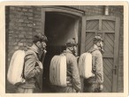 Drei Männer in Uniform mit Gasmaske und Rucksäcken vor einer offenen Tür eines Gebäudes.