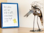 Vogelskulptur aus Metall neben einer  gerahmten Postkarte. Auf der Karte steht: "Das Leben wär' nur halb so nett, wenn keiner einen Vogel hätt"