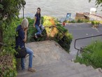 Eine Frau sitzt auf einem Treppengeländer, im Hintergrund der Rhein. Ein Mann steht oben auf der Treppe und fotografiert sie.