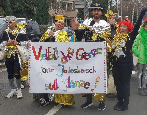 Eine Gruppe Kostümierter hält ein Banner hoch, auf dem steht: Vielfalt ohne Grenze janz Jodesbersch sull glänze.