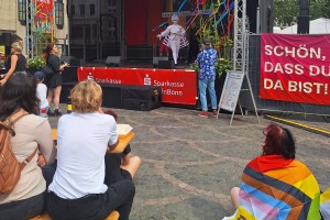 Auf einem Platz ist eine Bühne, dekoriert in Regenbogenfarben, aufgebaut, auf der eine Person in weißem Kostüm zu erkennen ist. Auf Bierbänken und dem Boden sitzen Zuschauer*innen. Eine Person hat eine Regenbogenflagge umgehängt.
