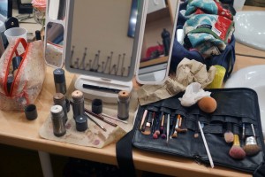 Auf einem Tisch ist eine Palette Make Up und Pinsel ausgebreitet, daneben steht ein Vergrößerungs-Spiegel.