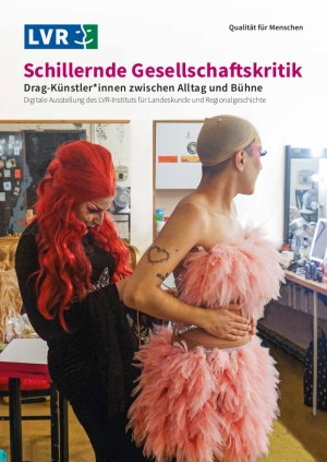 In einer Theatergarderobe steht eine Person mit Perückenkappe, die ein mit Federn besetztes rosafarbenes und silber-glitzerndes Kostüm trägt. Sie bekommt von einer zweiten Person in langer, roter Perücke und schwarzem Samtkleid, die hinter ihr steht, ein Korsett zugebunden.