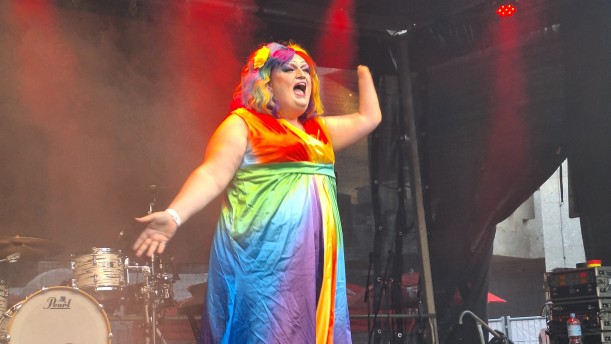 Eine Person mit langem Kleid, Perücke und Make-Up in Regenbogenfarben steht auf einer ausgeleuchteten Bühne, die Arme ausgebreitet, den Mund wie zum Singen weit geöffnet.