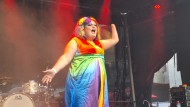 Eine Person mit langem Kleid, Perücke und Make-Up in Regenbogenfarben steht auf einer ausgeleuchteten Bühne, die Arme ausgebreitet, den Mund wie zum Singen weit geöffnet.