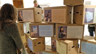 Eine Frau betrachtet eine Ausstellungsinstallation aus Umzugskartons auf denen Fotoporträts und Objektgeschichten abgebildet sind.