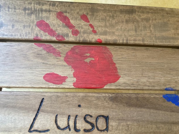 Auf einer Holzbank prangt der rote Handabdruck eines Kindes. Daneben steht der Name 'Luisa'.