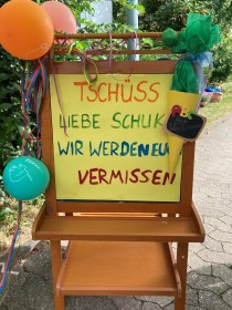 An einer Kindertafel hängt ein gelbes Plakat mit der bunten Aufschrift 'Tschüss liebe Schukis. Wir werden euch vermissen'. An das Plakat ist eine gebastelte gelbe Schultüte mit Tafel, ABC und 'Auf Wiedersehen' angeheftet.