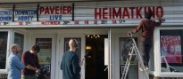 Foto: Ein Kamerateam filmt einen Mitarbeiter eines Kinos, welcher Buchstaben vor einem Kino aufhängt.
