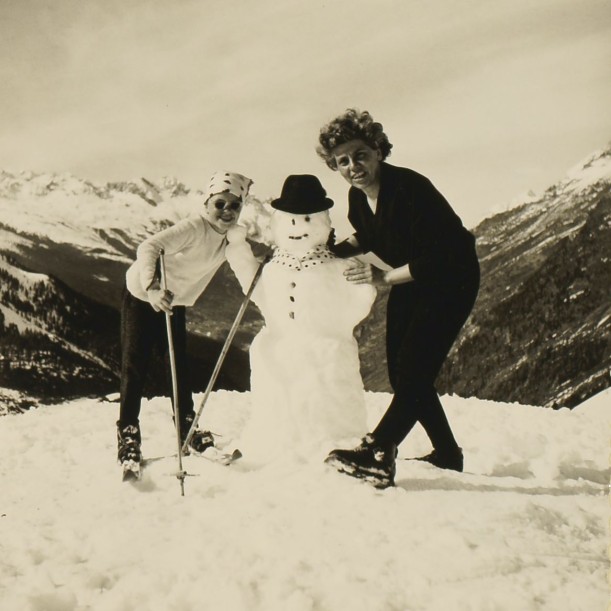 Auf einem Berg haben sich ein Kind mit Skiern und Skistöcken und eine Frau im Schnee für ein Foto positioniert. In ihrer Mitte steht ein Schneemann mit Hut. Einer der Skistöcke ist an ihn gelehnt. Das Kind und die Frau lachen in Richtung Kamera. Im Hintergrund erstreckt sich ein Bergpanorama.