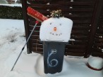 Aus einer hohen Schneeschicht auf einer Abfalltonne wurde der Kopf eines Schneemanns gestaltet, ein Straßenbesen ist gegen die Figur gelehnt. Hinter der Tonne ein Sichtschutzzaun, dahinter eine Hecke.