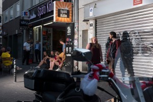 Straßenansicht eine Kioskes mit dem Namen "Späti", Junge Leute sitzen und stehen davor. Ein Mann unterhält sich mit dem Kioksinhaber.