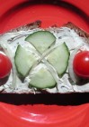Auf einem roten Teller liegt eine Scheibe Brot bestrichen mit Frischkäse und belegt mit Tomaten, einer Scheibe Gurke und Zwiebeln.