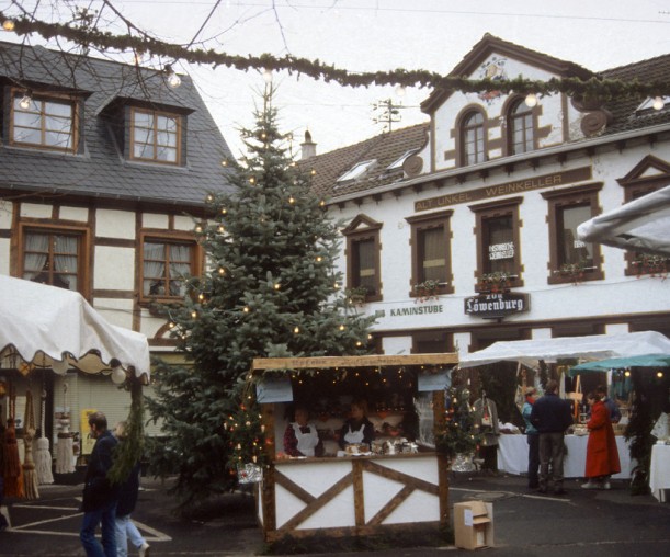 Ein kleiner Weihnachtsmarkt vor einer historischen Kulisse. Vor einer Häuserwand, hinter einem einfachen Stand, steht ein großer Weihnachtsbaum.