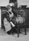 Eine Frau mit Kittelschürtze dreht an einer Kurbel die an einem kleinen Holzfass montiert ist