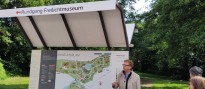 Ein Mann steht - redend vor einigen Zuhörenden - vor einer Tafel mit einer Grafik mit der Überschrift "Museumsplan". Darüber ein Dach mit der Aufschrift "Museumsrundgang"
