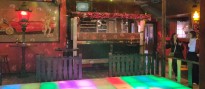 Innenraum einer Diskothek mit einem Fußboden aus bunt beleuchteten Kacheln, einer Diskokugel an der Decke und diversen Dekorationselementen im Hintergrund. Rechts neben der Tanzfläche stehen zwei Frauen.