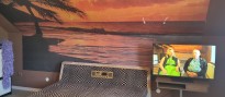 Breites Doppelbett vor einer großen Fototapete, die eine Palme vor einem Strand bei Sonnenuntergang oder Sonnennaufgang als Motiv hat. Neben dem Bett ein Bildschirm, das ein älteres Paar sitzend im gleichen Raum zeigt.