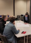 Um einen Tisch sitzen vier Personen. Eine stehende Frau beschriftet auf ein auf dem Tisch liegendes großes weißes Plakat etwas mit einem farbigen Stift.
