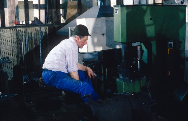 In einer Werkstatt sitzt ein Mann mit Hut und blauer Hose auf einem Hocker vor einer großen grün lackierten Maschine. Der Mann ist ein Schwertschmied beim Herstellen von Rohlingen für Säbel.