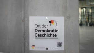 Weiße Tafel mit schwarzer Schrift und schwarz-rot-goldener Farbgebung auf grauem Hintergrund.