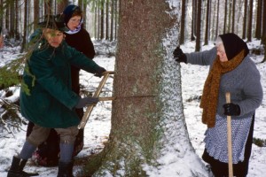 In einem verschneiten Wald stehen drei Personen um einen Baumstamm. Ein Mann in grüner Jacke und Hut (links) hält einen großen Messschieber an den Baum. Auf der anderen Seite des Baums (rechts) steht eine ältere Frau mit Schürze, braunem Schal und Kopftuch, die eine Axt in der Hand hält.