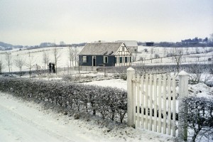 Altern. Bildtext: Ein allein stehendes Haus in einer schneebedeckten Landschaft. Vor dem Haus ist ein von einer Hecke umrahmter Garten erkennbar mit einem Tor aus weißen Holzlatten.