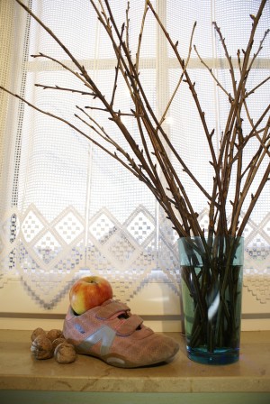Das Foto zeigt eine Glasvase mit abgeschnittenen Zweigen eines Obstbaumes auf einer Fensterbank. Links daneben steht ein Kinderschuh, der mit einem Apfel gefüllt ist. Neben dem Schuh liegen Walnüsse.