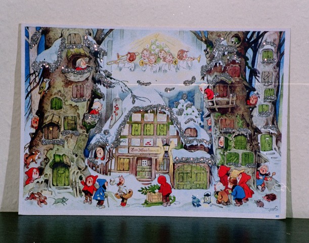 Klassischer Adventskalender mit Bildchen und weihnachtlichem Motiv: Eine Schneelandschaft mit zwei großen Bäumen, in denen kleine Heinzelmännchen leben. Am Himmel sind musizierende Engel zu sehen.