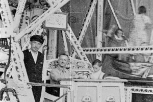 Die historische Schwarzweißfotografie zeigt eine Schiffschaukel in Betrieb mit Samtvorhängen, der verzierten Gerüstkonstruktion und einer Orgel hinter der Verkaufsloge.