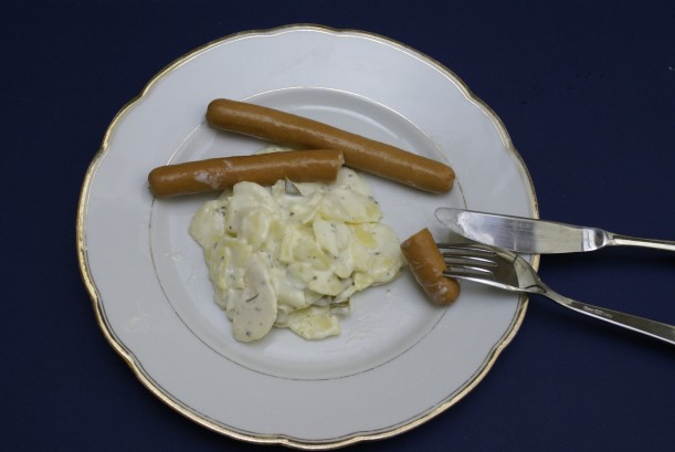 Goldrandteller mit Besteck auf dem eine Portion Kartoffelsalat sowie zwei Wiener Würstchen angerichtet sind. Ein Würstchen ist angeschnitten, das Stück Wurst steckt auf der Gabel.