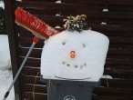 Auf einer Mülltonne liegt der Kopf eines Schneemanns, an den ein Besen angelehnt ist