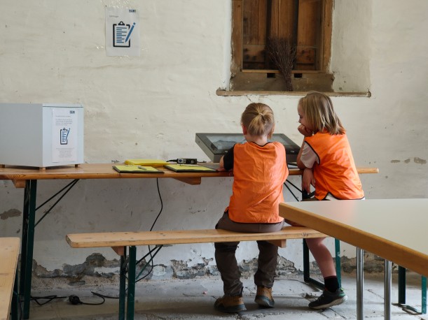 Zwei Kinder mit orangenen Westen sitzen auf einer Holzbank und betrachten einen großen Touch Screen.