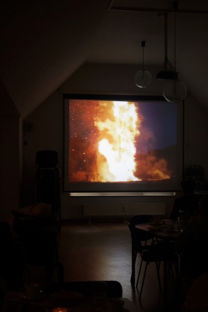 In einem dunklen Raum hängt eine Leinwand, auf der das Bild eines großen Feuers zu sehen ist. Unscharf sind im Vordergrund Tische und Stühle zu erkennen.