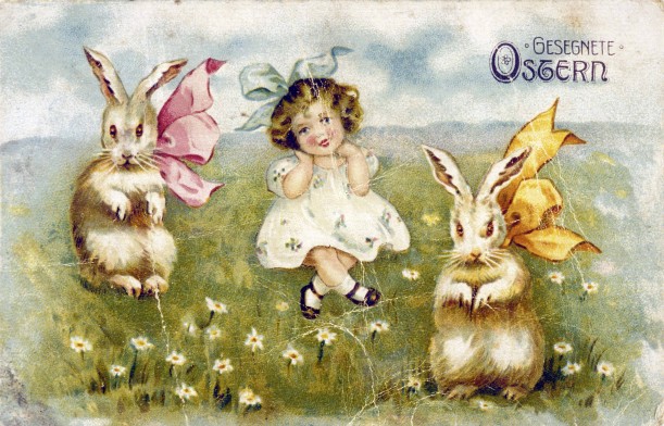 Ein Mädchen mit blauer Schleife im Haar sitzt zwischen zwei Osterhasen im grünen Gras.