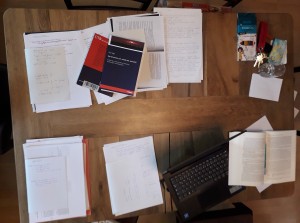 rechteckiger Holztisch auf dem sich Bücher, Zettelstapel, ein Laptop, ein Glas, Stifte und ein Schlüsselbund befinden