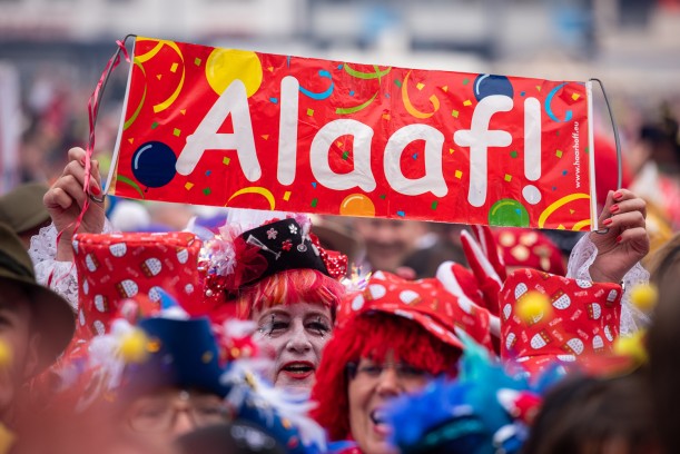 Eine Menschenmeng in bunten Kostümen, mit Hüten und Perücken hält ein Transparent hoch. Darauf steht: "Alaaf!"