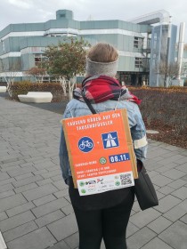 Rückenansicht einer Person auf einer Straße auf ihrem Rücksach ein Schild "Tausend Räder auf den Tausendfüssler" und die Einladung zu einer Fahrrad-Demo