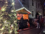 Straßenzug mit einer weihnachtlich beleuchteten Glühweinbude, an der mehrere Personen anstehen. Daneben ein Weihnachtsbaum
